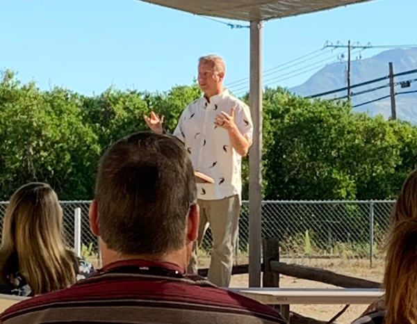 A speaker gestures while addressing an audience outdoors, surrounded by greenery and mountains in the background.