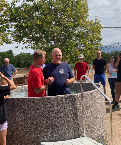 Two men stand in a baptismal font, surrounded by a group of people outdoors, with trees and cloudy sky in the background.