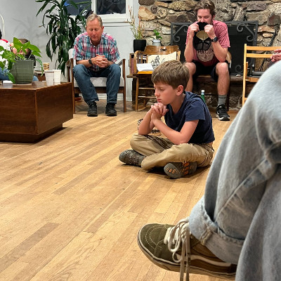 A young boy sits on the floor with hands clasped, while two adults are seated nearby in a cozy room.