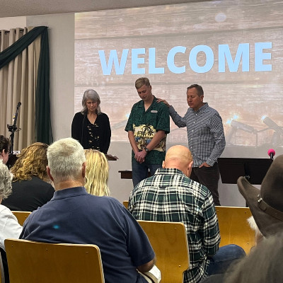 A group of three people stand together on stage; the word "WELCOME" is displayed prominently in the background.