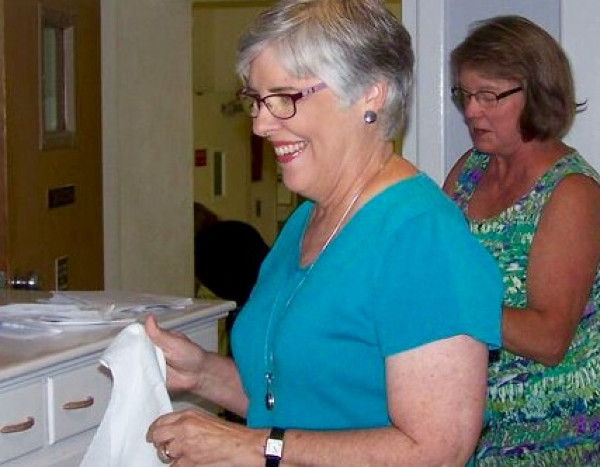 Two women in a bright room, one smiling and holding a cloth, the other in a patterned dress, engaged in an activity.