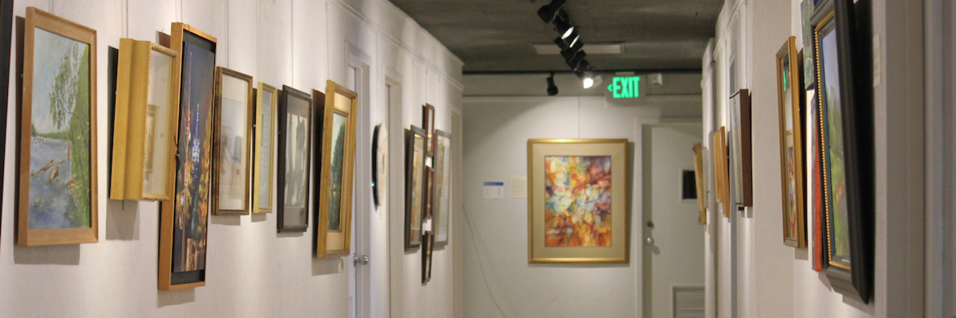 A hallway displaying various framed artworks, with an exit sign visible at the end.