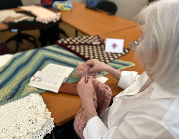 An elderly woman knitting with various colorful textiles and a book open on the table in front of her.