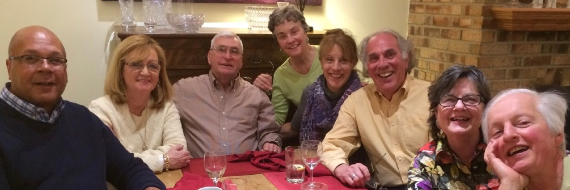 A group of eight people smiling together at a table with drinks, colorful tablecloth, and a cozy setting.