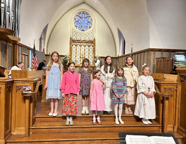 Eight girls in colorful dresses sing in a church, with stained glass windows and a wooden altar in the background.