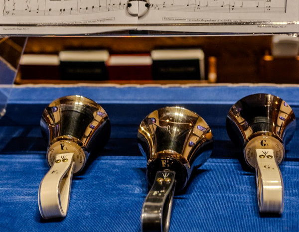 Three handbells labeled F, E, and G on a blue tablecloth, with sheet music visible above.