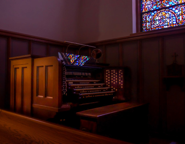 Wooden church organ with colorful stained glass window in background, soft lighting, and dark wooden paneling.