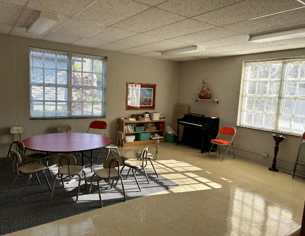 Bright classroom with a round table, chairs, shelves, and a piano; large windows letting in natural light.
