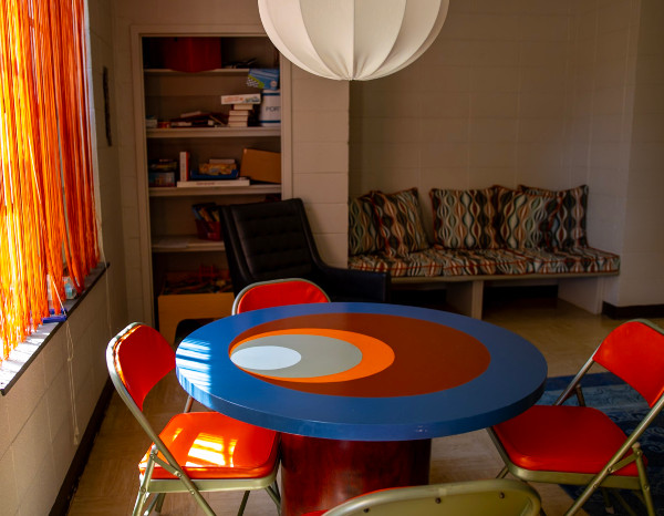 Colorful round table with concentric circles, surrounded by four red chairs, in a cozy room with a patterned couch.