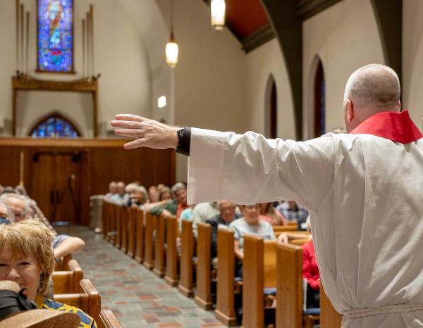 A man in a white robe speaks at a pulpit with a decorative cloth, stained glass window in the background.