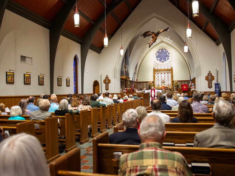 Interior of a church with a colorful ceiling, stained glass window, and a decorative wooden feature hanging above the altar.