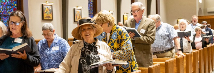 A group of people singing in a church, holding hymn books, with stained glass windows in the background.