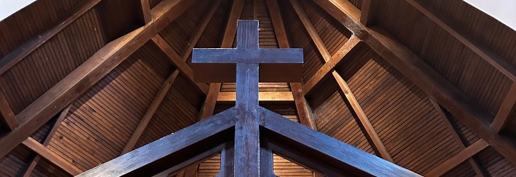 Interior view of a wooden church ceiling with a large cross at the center, showcasing a triangular architecture.