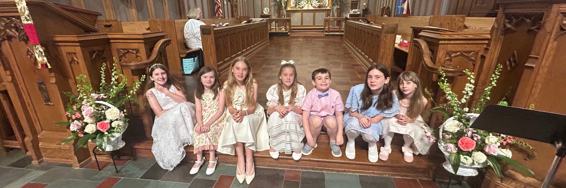 A group of seven children in formal attire seated on steps in a church, with floral arrangements nearby.