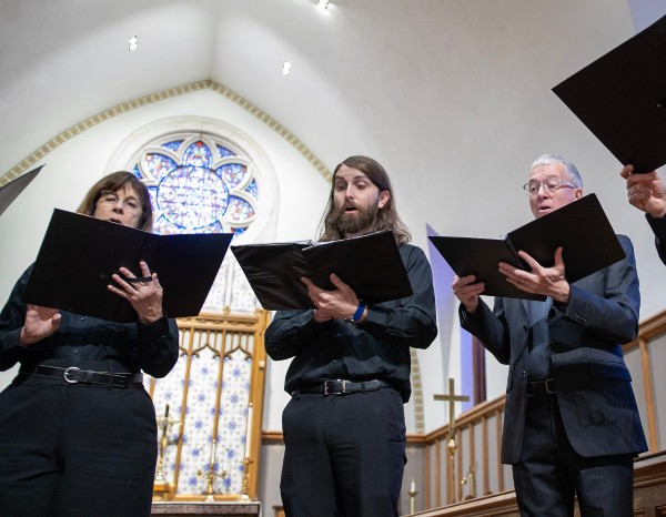 A choir sings in a church, holding music sheets, with a stained glass window in the background.