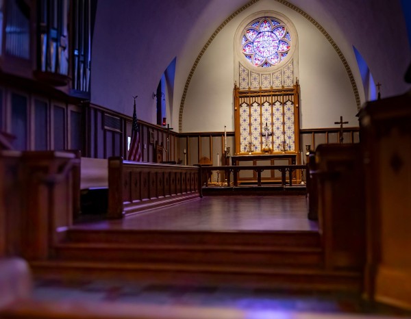 Interior of a church with a wooden altar, stained glass window, and pews in soft lighting.