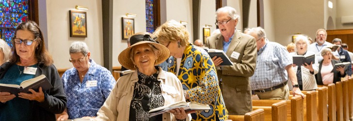 A group of people singing in a church, holding hymn books, with stained glass windows in the background.