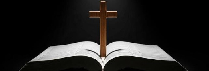 An open Bible with a wooden cross placed upright in the center, set against a dark background.