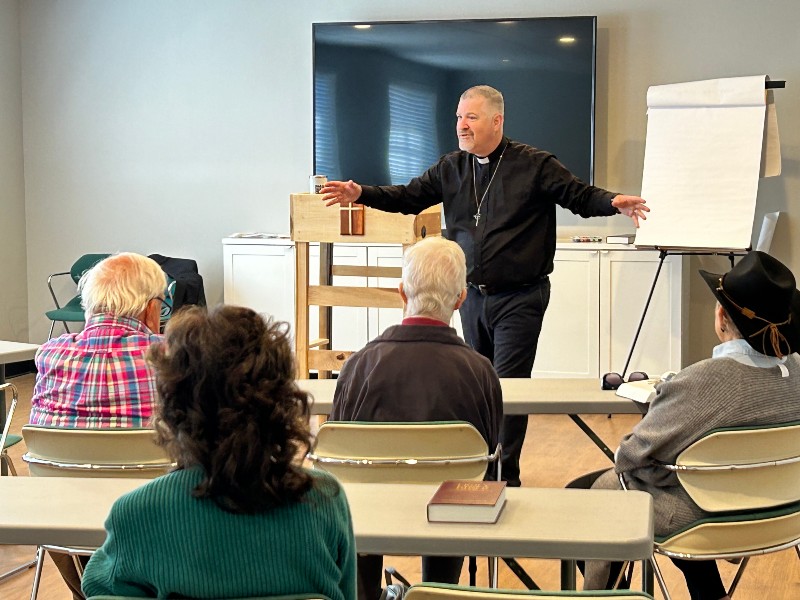 A speaker gestures while presenting to a small group seated at tables in a bright room. A whiteboard is visible.