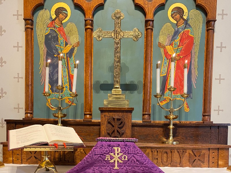 Altar with two angel icons, a cross, candles, and an open book, draped in purple cloth with "A P O" symbol.