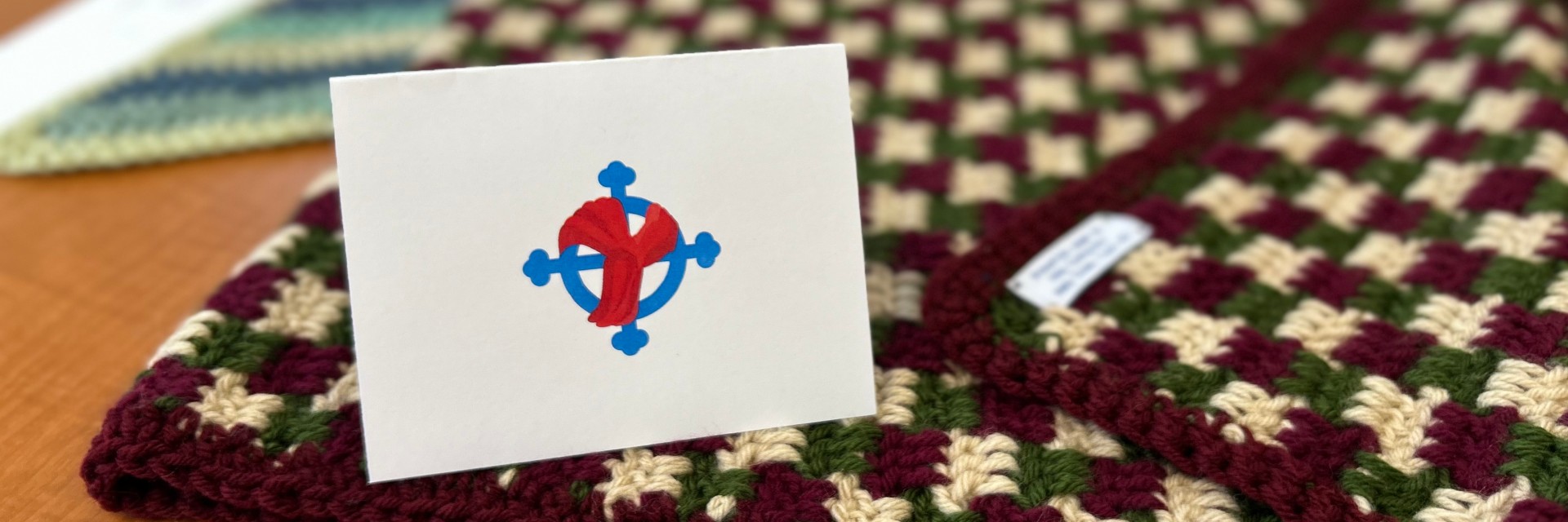 A crocheted blanket in red, green, and cream with a card featuring a blue and red design on a white background.